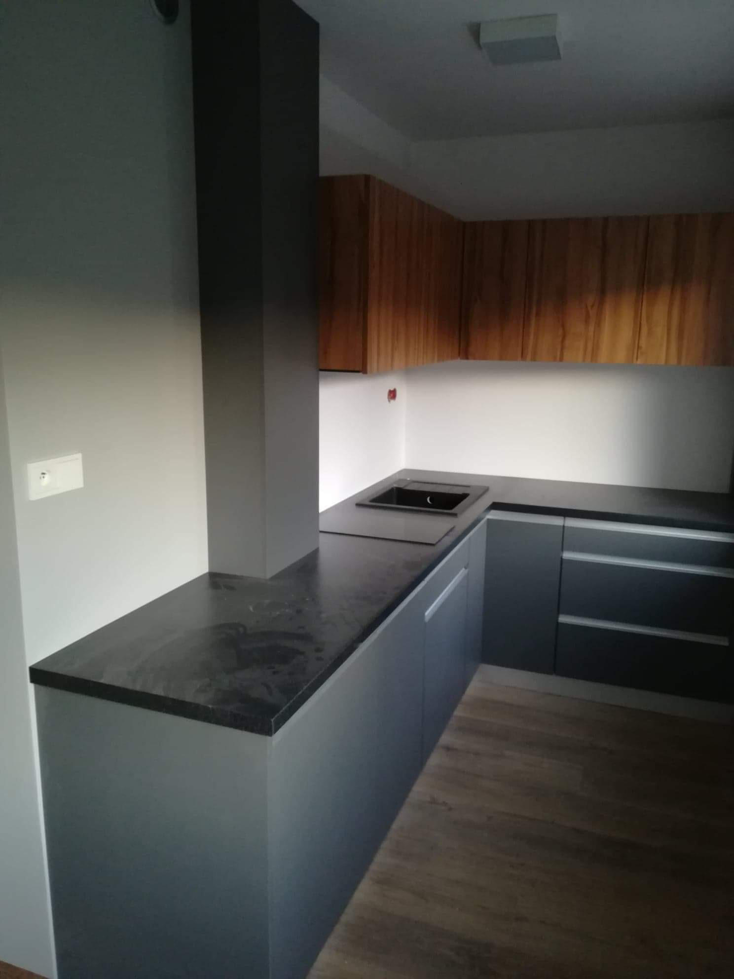 Nowoczesna kuchnia z szarymi szafkami, czarnym blatem i drewnianymi szafkami górnymi, z widocznym fragmentem drewnianej podłogi.