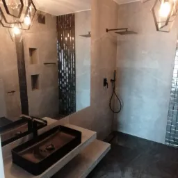 Nowoczesna łazienka z szarymi płytkami, czarną umywalką nablatową i geometrycznymi lampami wiszącymi, widoczne elementy armatury prysznicowej w kolorze czarnym.