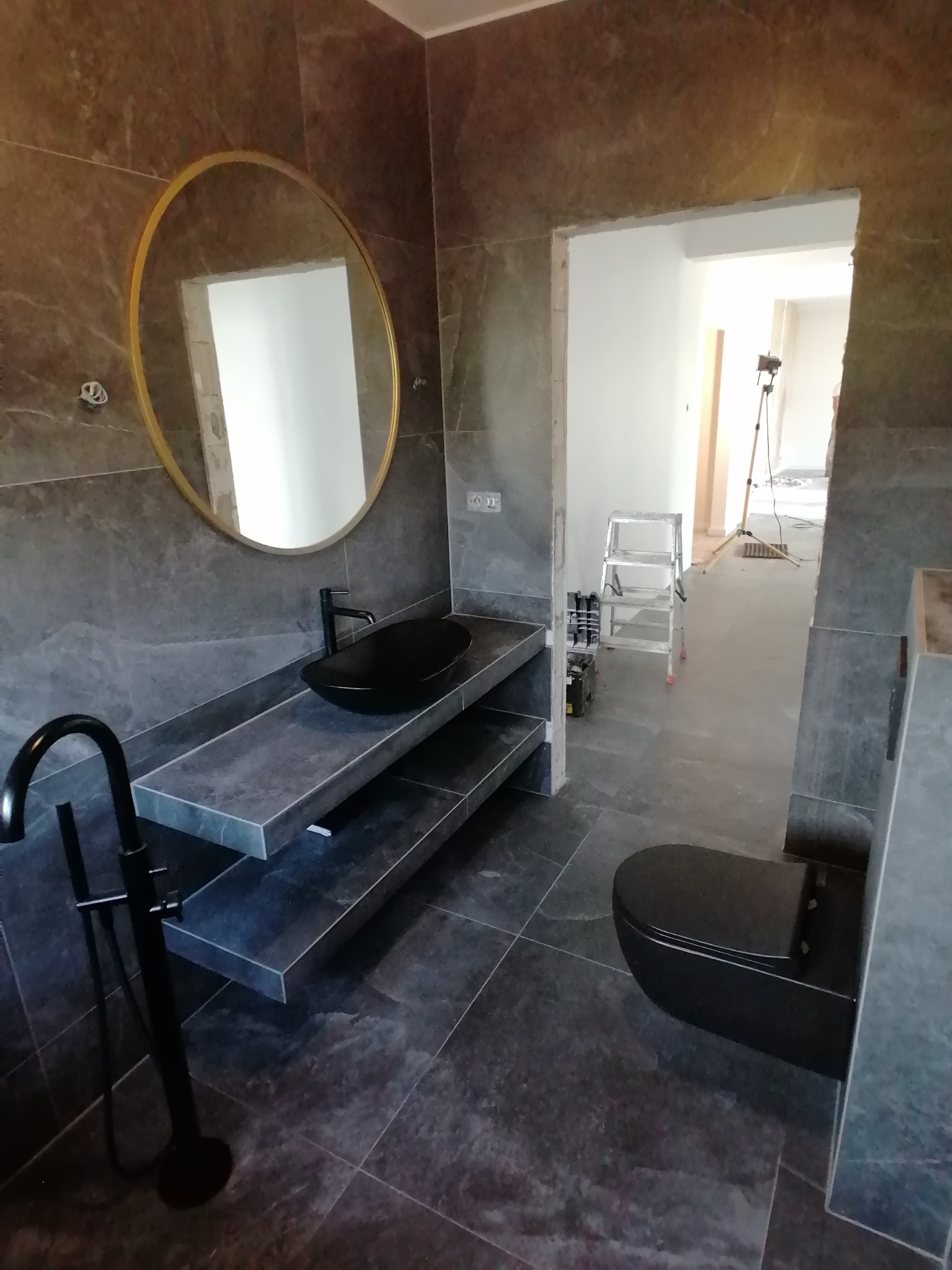 Nowoczesna łazienka w trakcie remontu: ciemne płytki imitujące kamień, czarna umywalka nablatowa i toaleta, złote okrągłe lustro, widoczny drabina i narzędzia w sąsiednim pomieszczeniu.