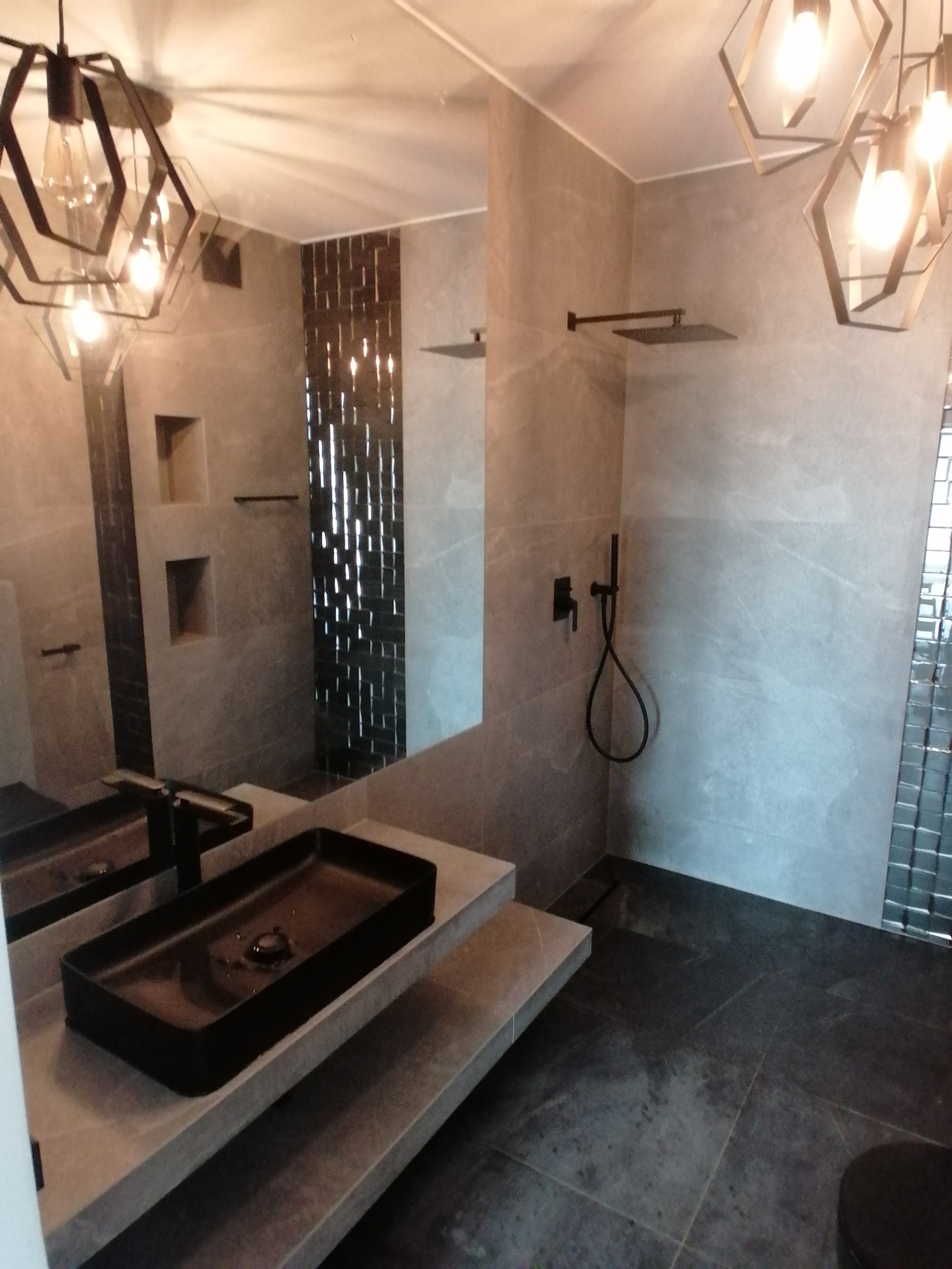 Nowoczesna łazienka z szarymi płytkami, czarną umywalką nablatową i geometrycznymi lampami wiszącymi, widoczne elementy armatury prysznicowej w kolorze czarnym.