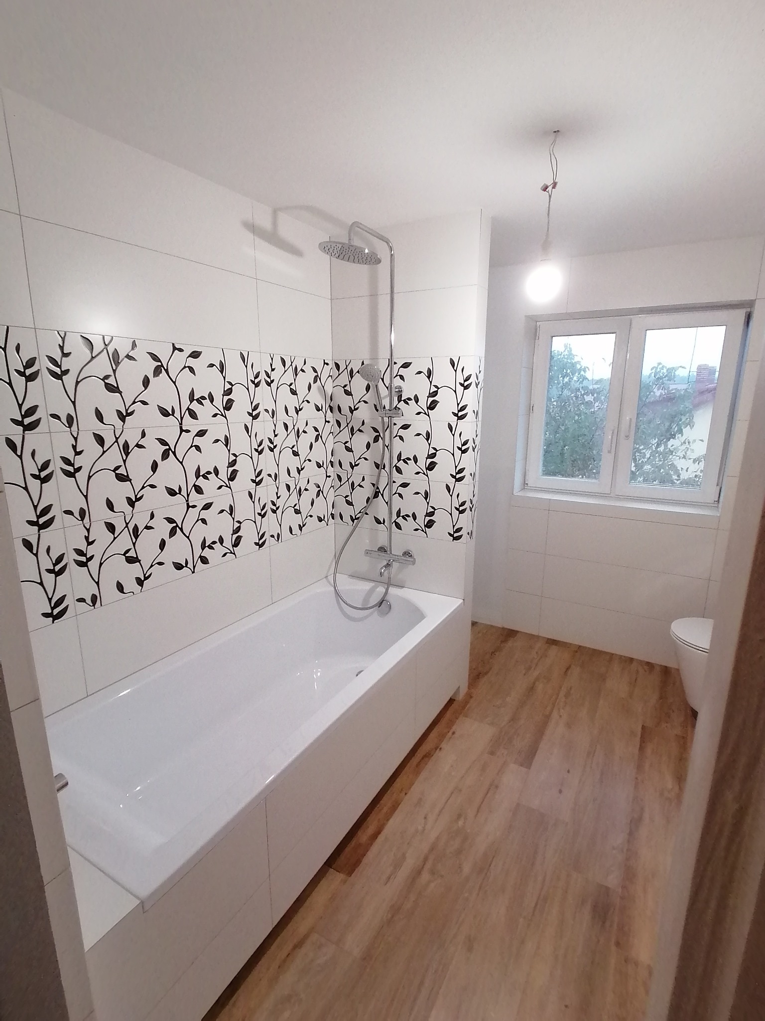 Wykończona łazienka w Sokołowie Podlaskim z wanną obudowaną białymi płytkami, nad nią dekoracyjne płytki z motywem roślinnym, drewnopodobna podłoga, widok na okno i częściowo widoczna toaleta.