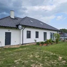 Jednopiętrowy dom z białą elewacją, ciemną stolarką okienną i grafitowym dachem, widok z ogrodu w pochmurny dzień, na elewacji widoczna antena satelitarna.