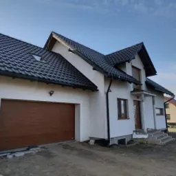 Biały dom jednorodzinny z brązową bramą garażową i ciemną dachówką, widok z zewnątrz w słoneczny dzień, teren wokół domu w trakcie prac wykończeniowych.