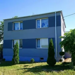 Dwukondygnacyjny budynek pomalowany w odcieniach szarości i granatu z widocznymi oknami, otoczony zielonym trawnikiem i drzewami w słoneczny dzień.