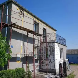 Dom w trakcie ocieplania elewacji: widoczne rusztowania, częściowo ocieplona ściana z białym styropianem, pianka montażowa w szczelinach, niebieski balkon z ozdobną balustradą i budynek gospodarczy...
