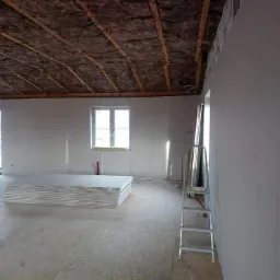 Wnętrze w trakcie prac wykończeniowych: ocieplony strop, ściany obłożone płytami karton-gips, stos płyt na podłodze, drabina oparta o ścianę.
