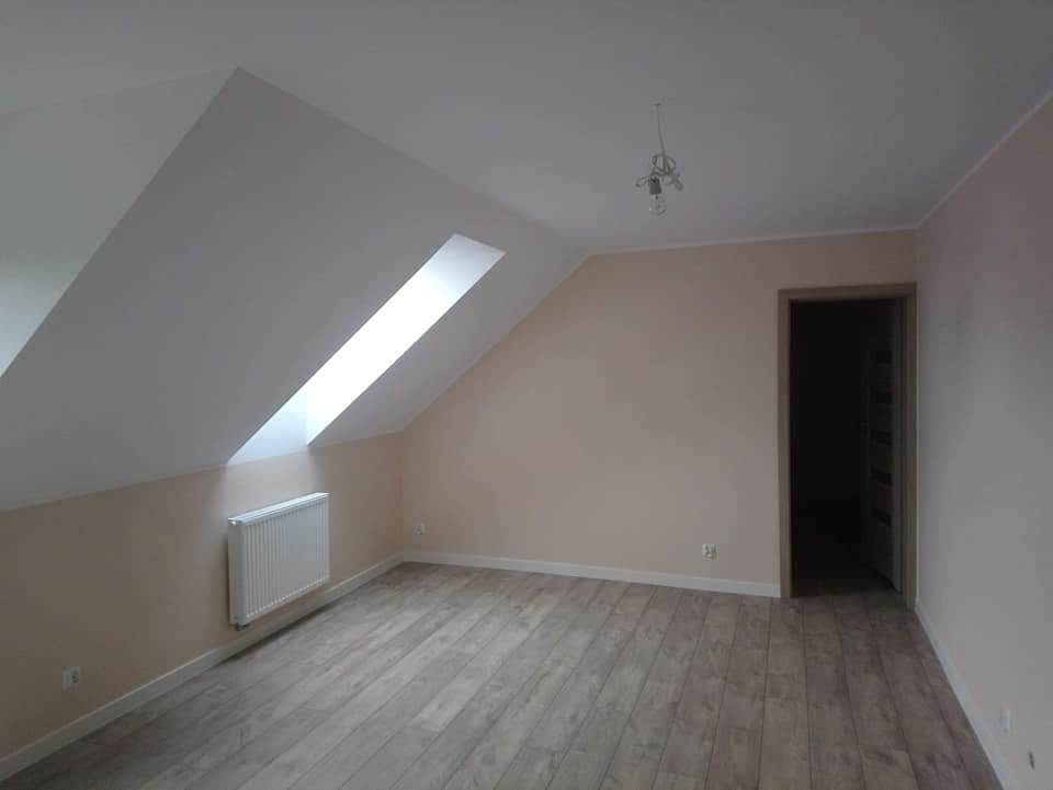 Poddasze po remoncie: skośny sufit z oknem dachowym, jasne ściany w odcieniu różu, drewnopodobna podłoga, biały grzejnik na ścianie, widoczne gniazdko elektryczne, drzwi bez ościeżnicy w głębi pokoju.