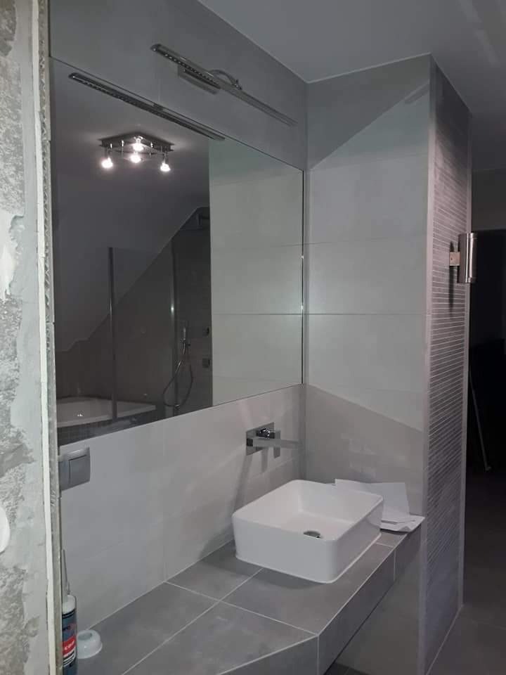 Nowoczesna łazienka w odcieniach szarości z kwadratową umywalką nablatową, chromowaną baterią i dużym lustrem na ścianie z szarych płytek. W lustrze odbija się wanna i oświetlenie sufitowe.