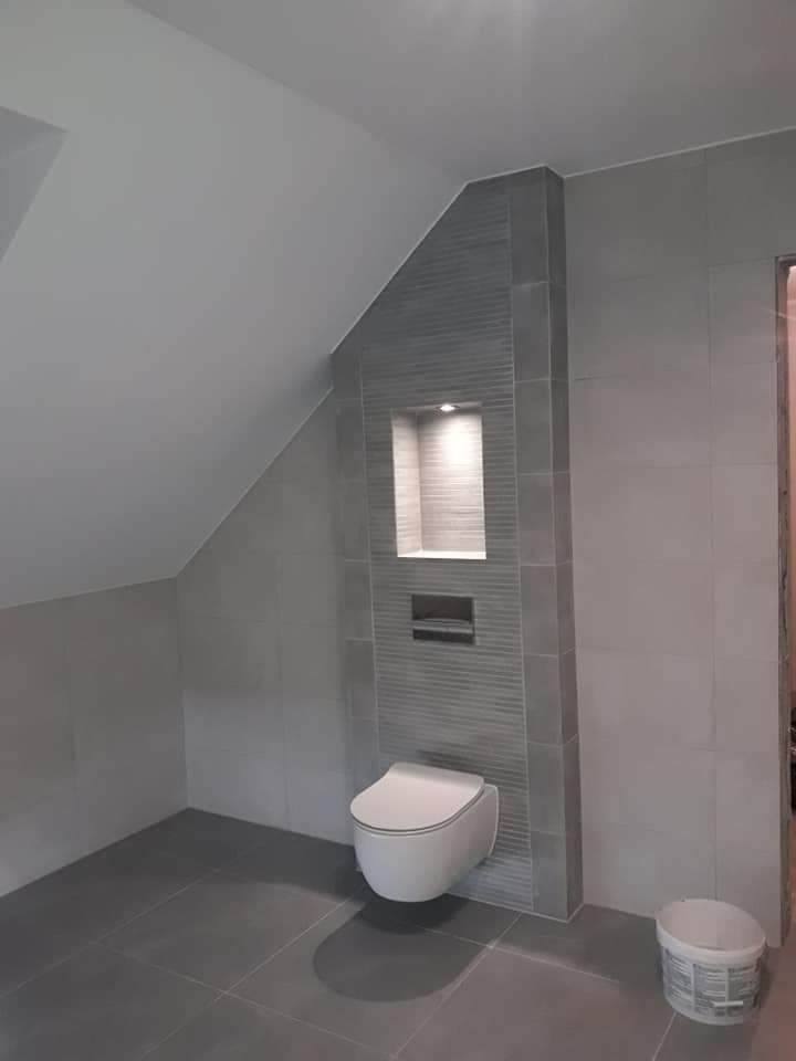 Szara łazienka na poddaszu z podwieszaną toaletą, wbudowaną półką z oświetleniem i wiaderkiem po farbie w rogu.