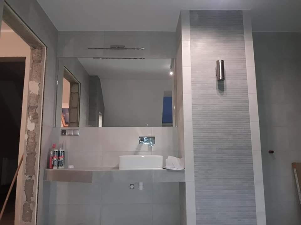 Szara łazienka w trakcie remontu, widoczne płytki, umywalka, lustro i niedokończone obramowanie drzwi z odsłoniętym murem.