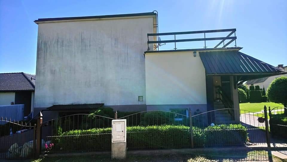 Dwupiętrowy dom z widocznymi śladami zabrudzeń i zacieków na elewacji, z balkonem na drugim piętrze i metalową bramą w tle zielonego trawnika.