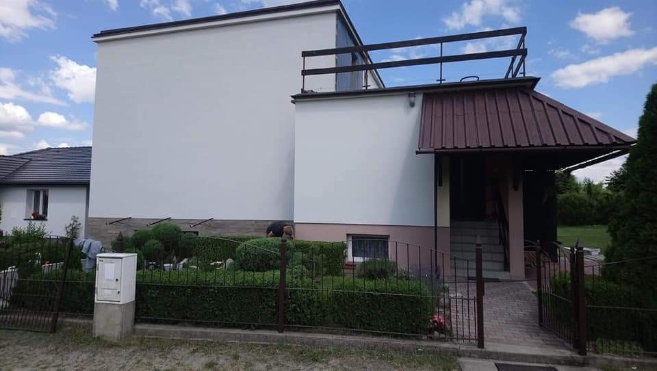 Dwukondygnacyjny dom po malowaniu elewacji na kolor biały, z widocznym fragmentem ogrodzenia i zadbanego ogrodu z krzewami, na tle błękitnego nieba z chmurami.