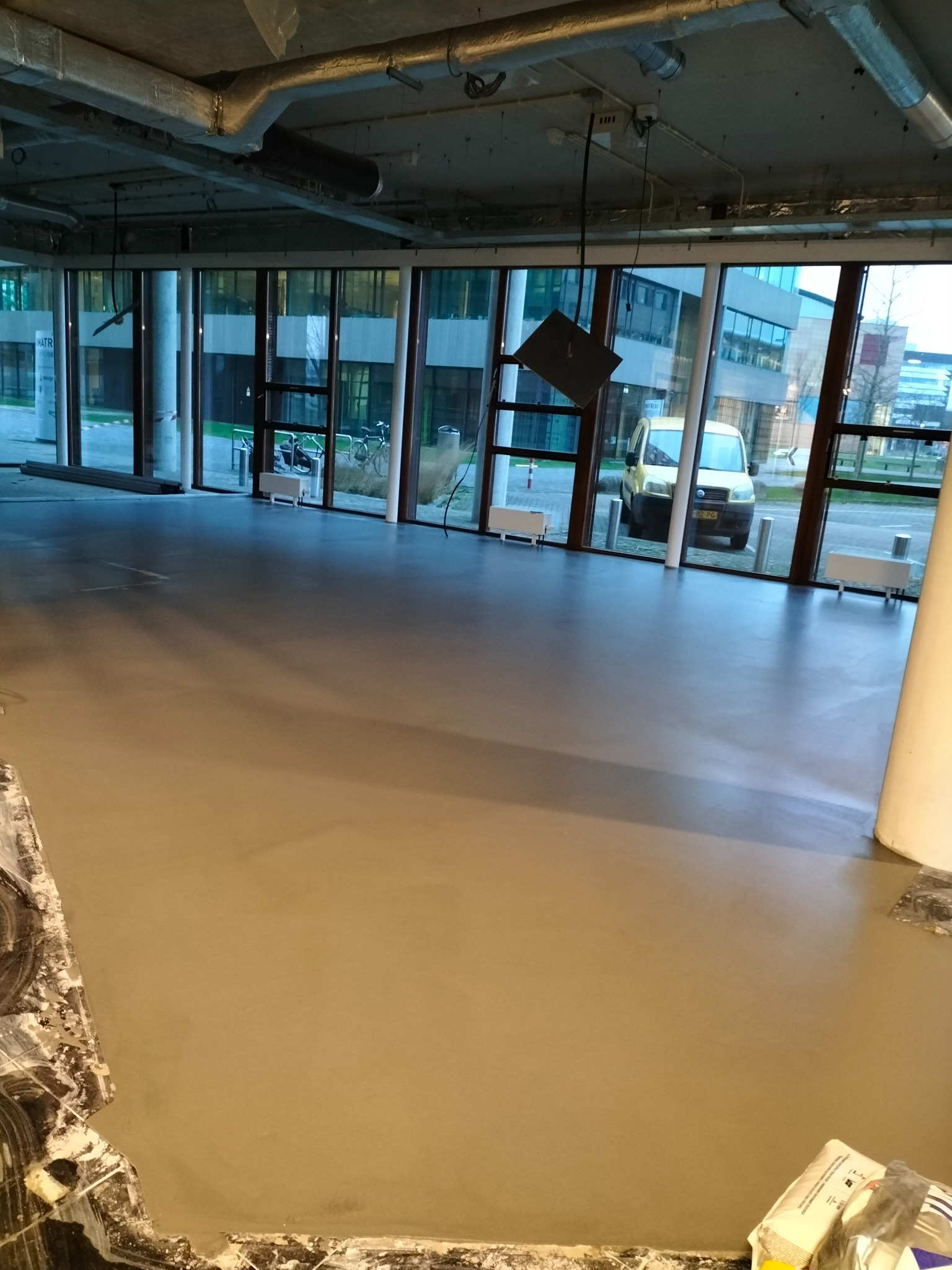 Świeżo wylana, gładka posadzka w przestronnym pomieszczeniu biurowym z widokiem na parking i zaparkowany samochód dostawczy za oknem.