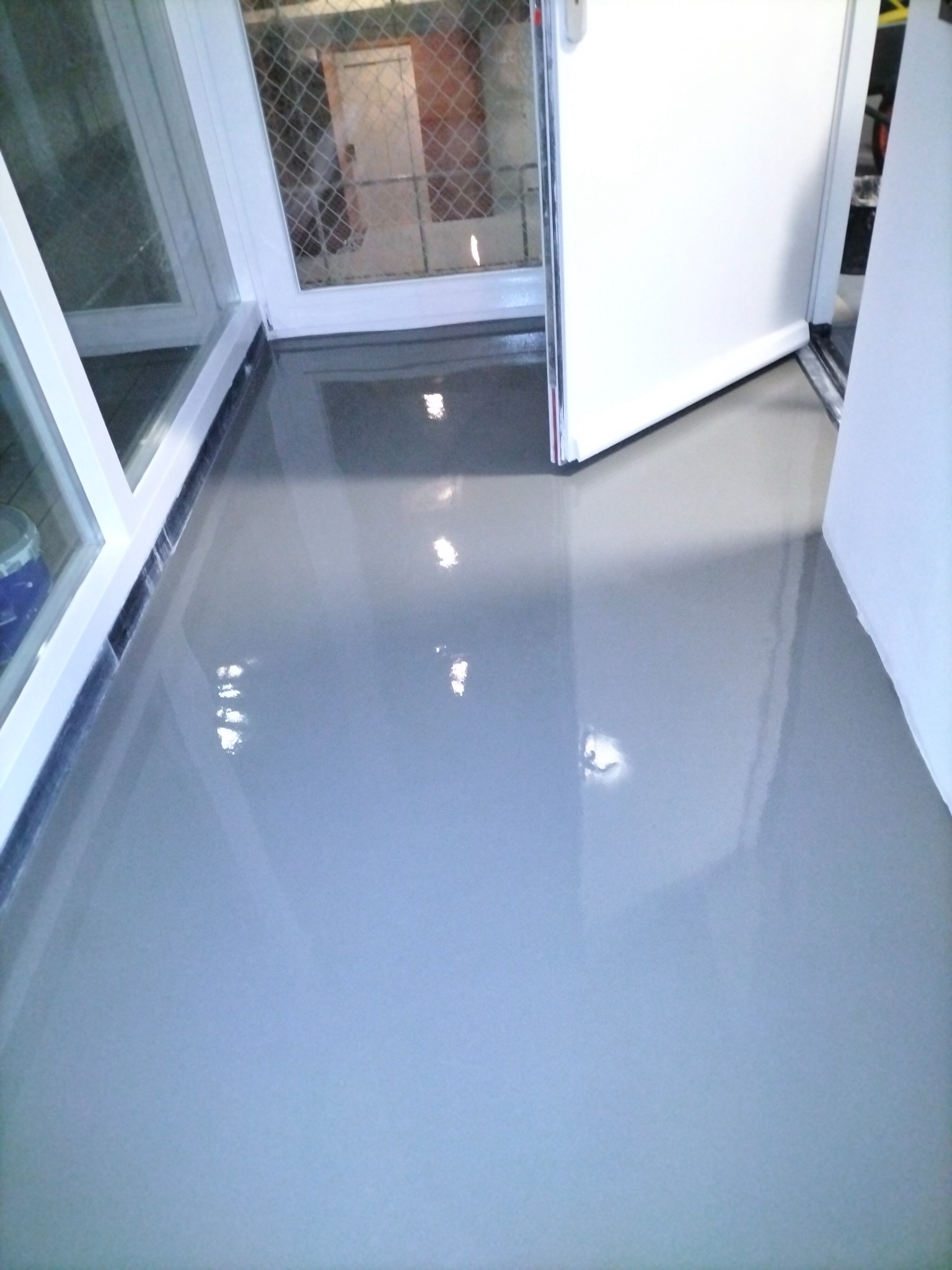 Świeżo wylana, gładka, szara posadzka z widocznymi refleksami światła, biała rama okienna i częściowo otwarte białe drzwi w tle.