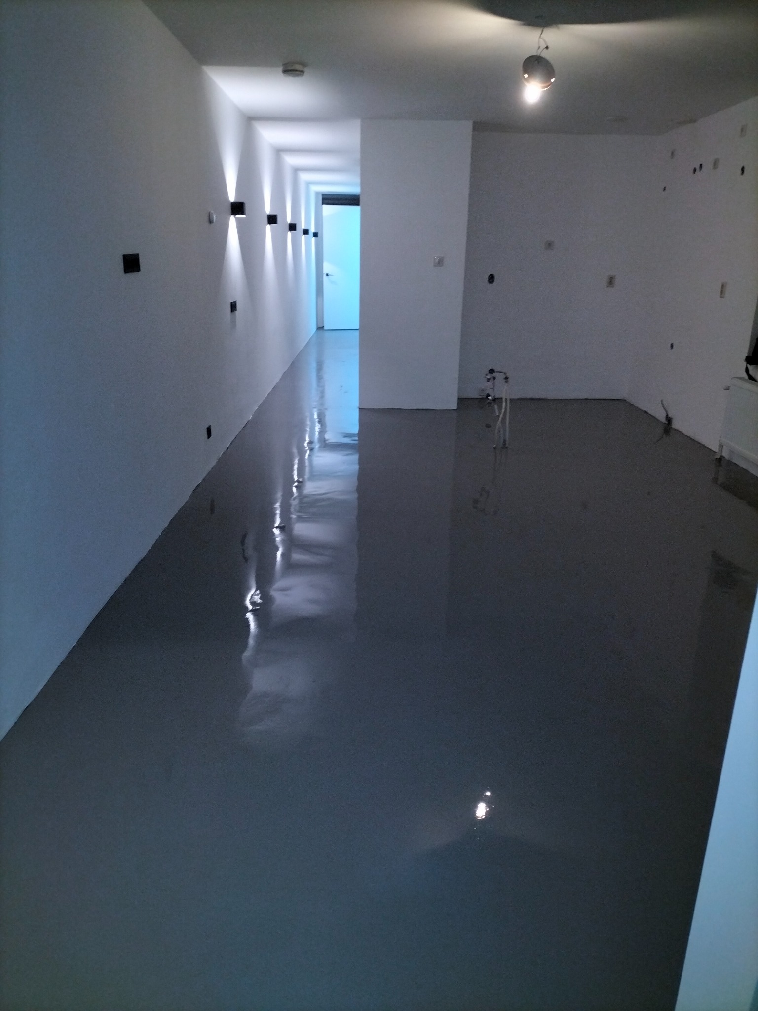 Świeżo wylana, szara posadzka jastrychowa w długim korytarzu z białymi ścianami i nowoczesnym oświetleniem ściennym, widoczne instalacje hydrauliczne w tle.