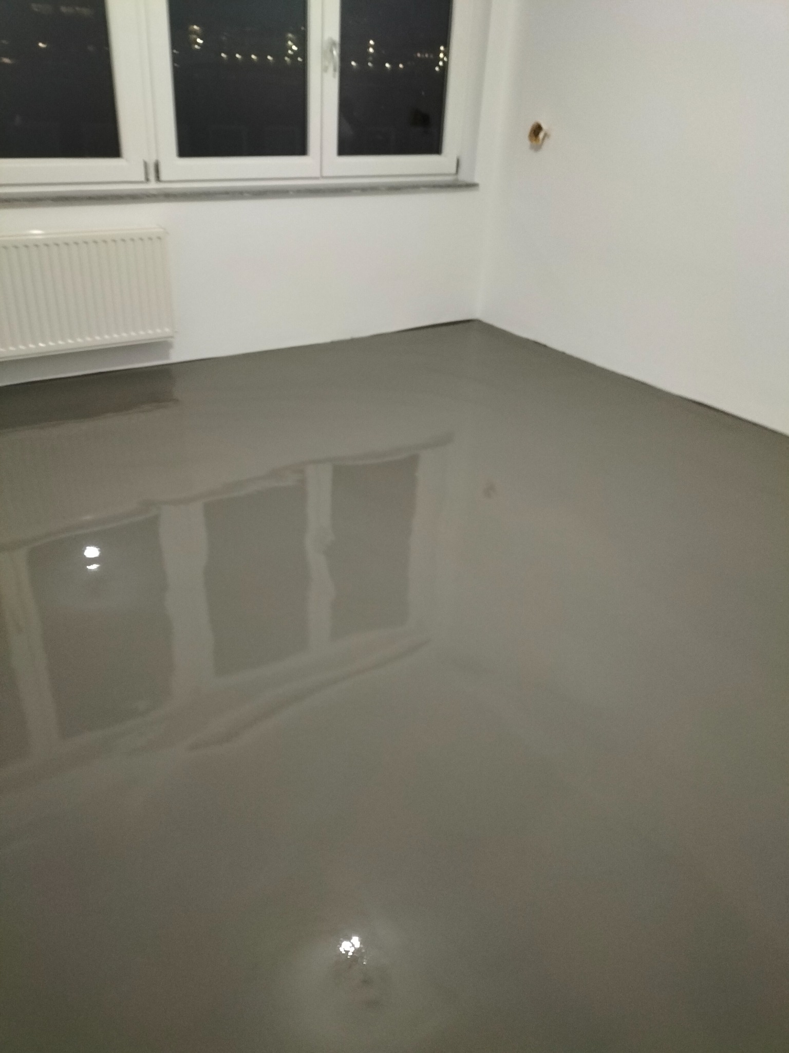 Świeżo wylana, szara, samopoziomująca posadzka w pomieszczeniu z białymi ścianami, grzejnikiem i oknem z widokiem na nocne miasto.