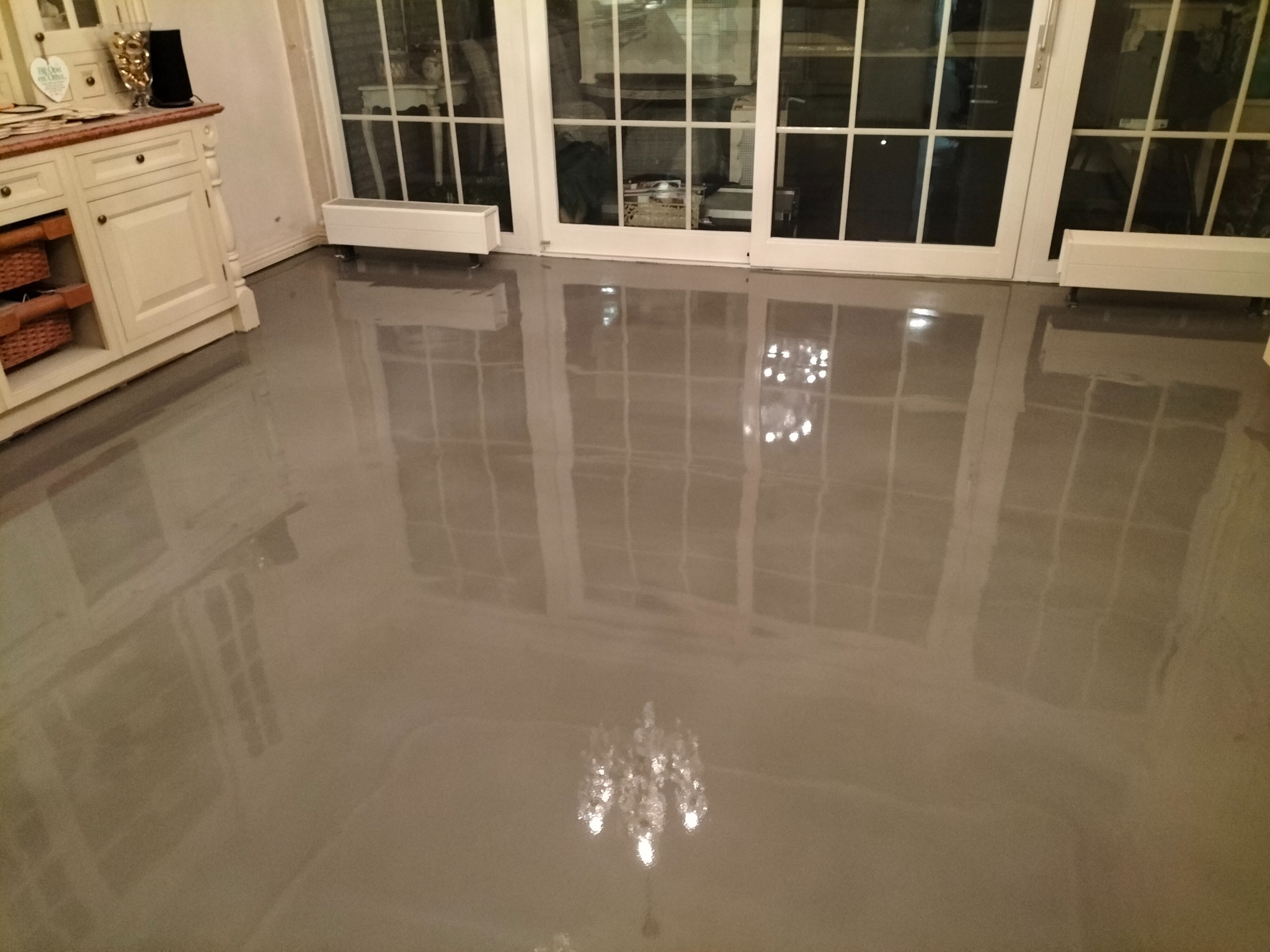 Błyszcząca, szara posadzka w jasnym pomieszczeniu z białymi meblami i dużymi oknami balkonowymi, odbijająca światło żyrandola.