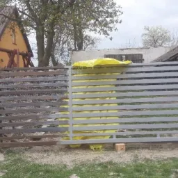 Połowa metalowej bramy po piaskowaniu i przed malowaniem proszkowym, druga połowa w trakcie piaskowania, zabezpieczona żółtą folią, widoczne budynki w tle.