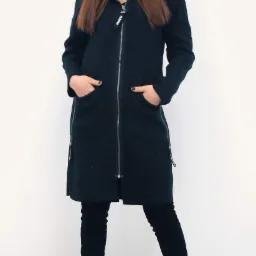 Modelka w długim, grafitowym płaszczu z kapturem, zapinanym na zamek błyskawiczny, z rękami w kieszeniach, na białym tle.