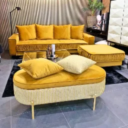 Komplet wypoczynkowy w odcieniach musztardowej żółci: sofa z poduchami, pufa i podnóżek z wzorem w pepitkę na złotych nóżkach, ustawione na ciemnoszarym dywanie, z dekoracyjnym stolikiem i lampą...