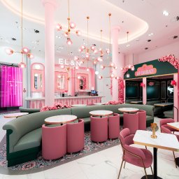 ZABŁYSZCZ Damian Błyszcz - Wnętrze kawiarni w stylu art deco z tapicerowanymi, różowymi krzesłami i zieloną, półokrągłą sofą, dekorowane różowymi kwiatami i nowoczesnymi lampami wiszącymi.