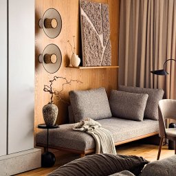 ZABŁYSZCZ Damian Błyszcz - Szary tapicerowany szezlong z poduszkami i pledem w nowoczesnym wnętrzu z drewnianą boazerią, dekoracjami i oświetleniem.