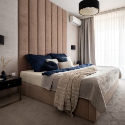 ZABŁYSZCZ Damian Błyszcz - Sypialnia z łóżkiem kontynentalnym, wezgłowiem z paneli tapicerowanych w odcieniach beżu, ciemnoniebieskimi poduszkami i beżową narzutą, obok czarna szafka nocna z wazonem.