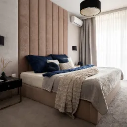 Sypialnia z łóżkiem kontynentalnym, wezgłowiem z paneli tapicerowanych w odcieniach beżu, ciemnoniebieskimi poduszkami i beżową narzutą, obok czarna szafka nocna z wazonem.