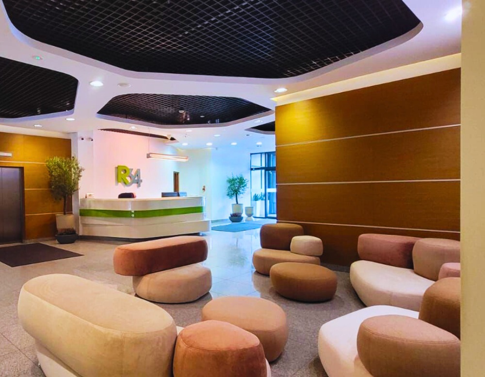 Nowoczesne lobby biurowca z tapicerowanymi pufami w odcieniach beżu i brązu, recepcją z logo R4 i roślinami w donicach.
