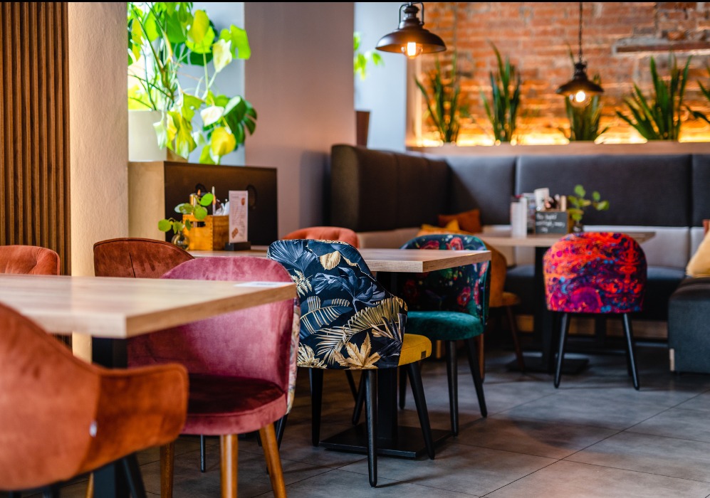 Wnętrze kawiarni KATOSIX6 z różnorodnymi krzesłami tapicerowanymi: aksamitne w odcieniach różu i pomarańczy, krzesło z motywem liści palmowych na granatowym tle, oraz pufa w intensywnych kolorach.