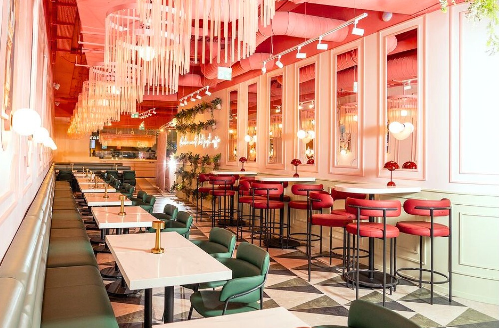 Wnętrze restauracji w Opolu z długą, zieloną tapicerowaną ławą, czerwonymi krzesłami barowymi i białymi stolikami, oświetlone dekoracyjnymi lampami i wiszącymi instalacjami.