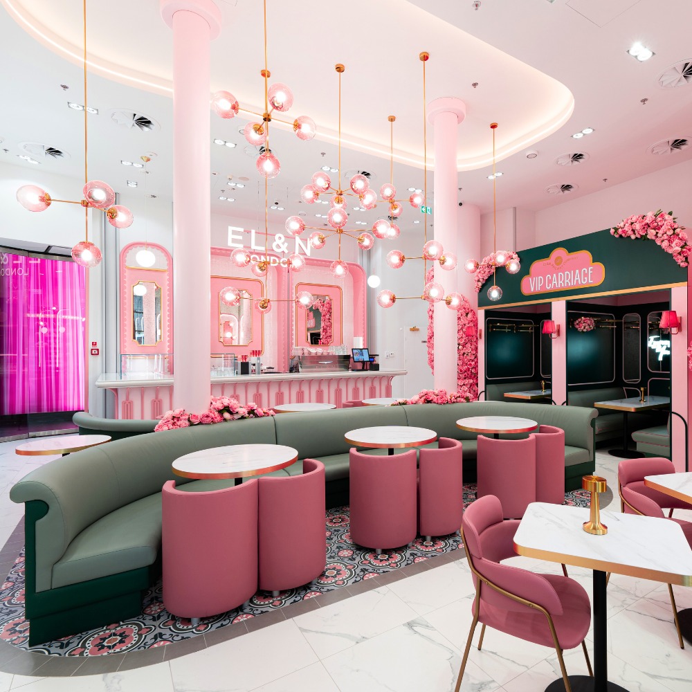 Wnętrze kawiarni w stylu art deco z tapicerowanymi, różowymi krzesłami i zieloną, półokrągłą sofą, dekorowane różowymi kwiatami i nowoczesnymi lampami wiszącymi.