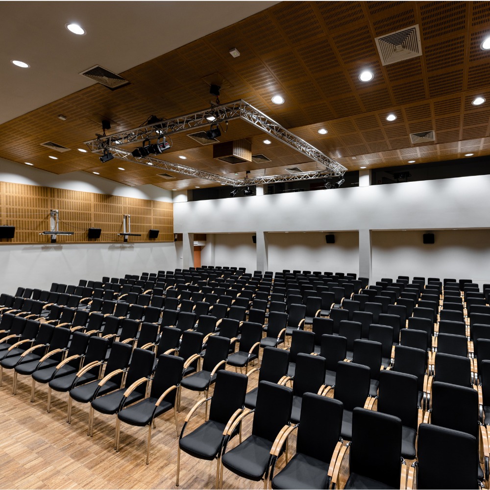 Sala konferencyjna z rzędami czarnych krzeseł z drewnianymi podłokietnikami, oświetlona metalową konstrukcją z reflektorami pod drewnianym sufitem z kratką.