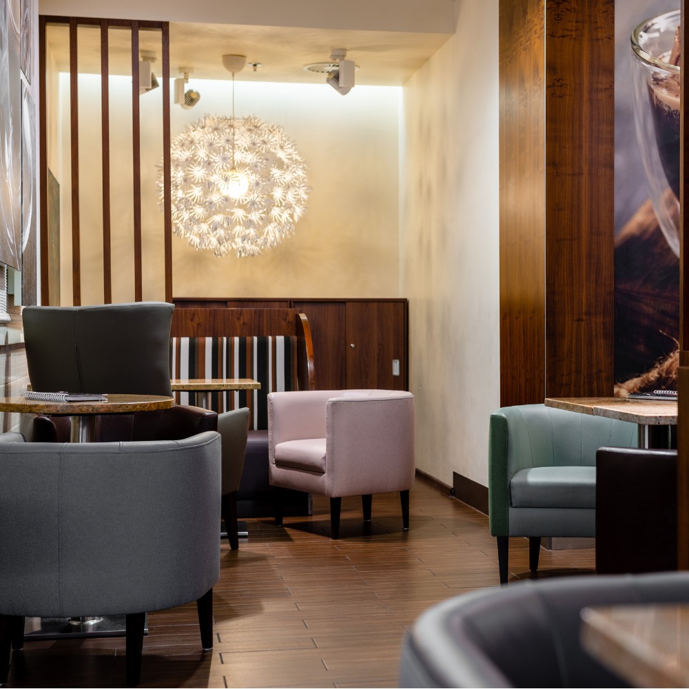 Wnętrze kawiarni z różnymi fotelami tapicerowanymi: szarymi, różowym i miętowym, drewniane panele na ścianach i dekoracyjna lampa wisząca w kształcie kuli.