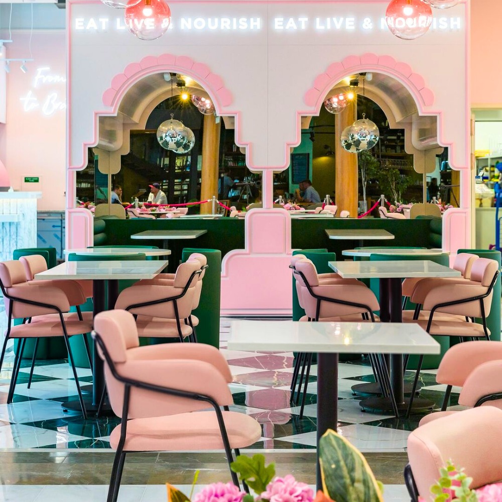 Różowe krzesła z czarnymi nogami i zielone tapicerowane loże w restauracji z neonowym napisem 'EAT LIVE & NOURISH' i podłogą w czarno-białą szachownicę.