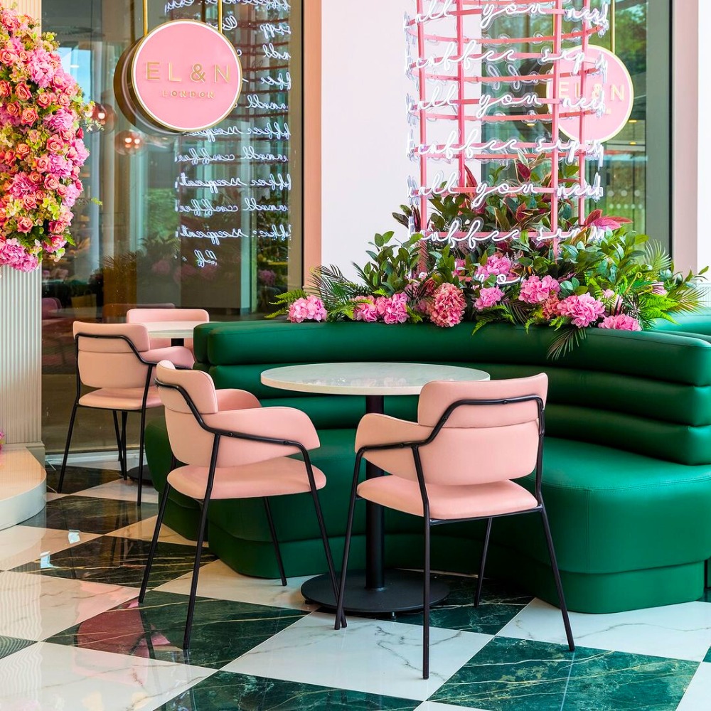 Różowe krzesła tapicerowane z czarnymi nogami przy okrągłym stoliku i zielonej kanapie w kawiarni EL&N London, dekoracja z różowych kwiatów i neonów.