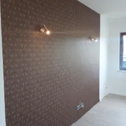 Ściana pokryta brązową tapetą w geometryczny wzór, oświetlona dwoma złotymi kinkietami, z widocznym gniazdkiem elektrycznym i fragmentem okna w tle.