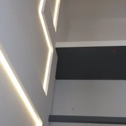 Nowoczesne oświetlenie sufitowe z liniowymi lampami LED wpuszczonymi w białą płytę kartonowo-gipsową, z fragmentem pomalowanego na szaro sufitu w tle.