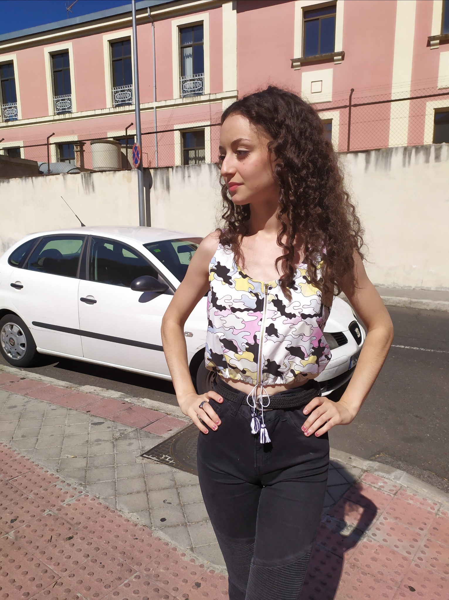 Młoda kobieta w krótkim topie z zamkiem i wzorem w pastelowe plamy, czarnych spodniach i plecionym pasku, pozuje na tle białego samochodu i różowego budynku.
