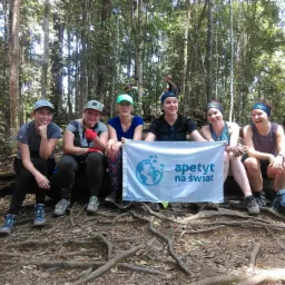 Grupa sześciu kobiet siedzi na korzeniach drzewa w lesie, trzymając biały baner z logo 'apetyt na świat'.