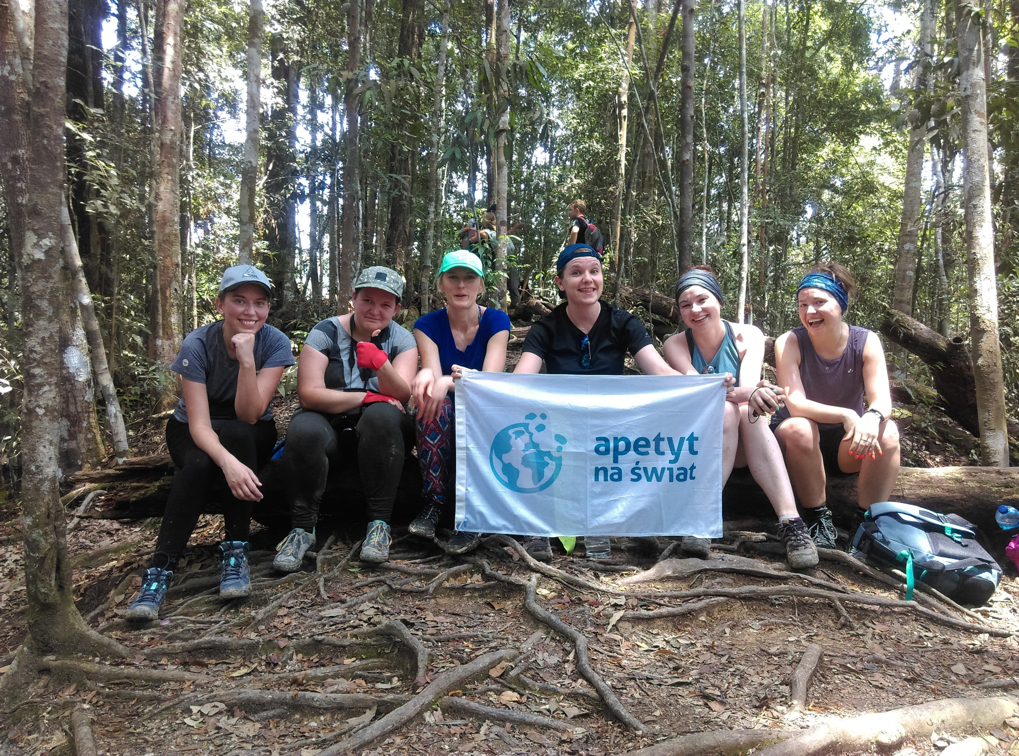 Grupa sześciu kobiet siedzi na korzeniach drzewa w lesie, trzymając biały baner z logo 'apetyt na świat'.