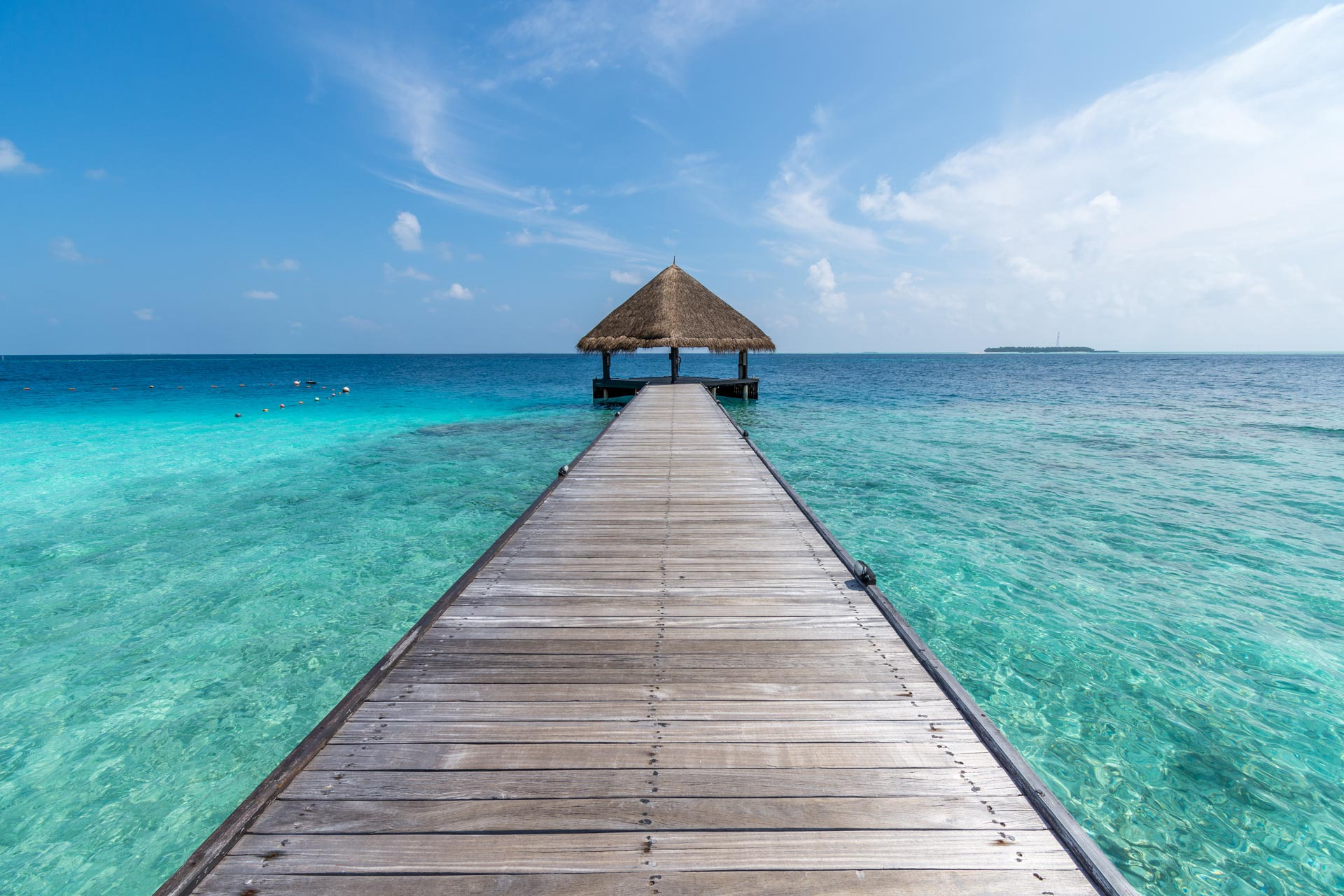 Długa drewniana kładka prowadząca do altany z trzcinowym dachem na turkusowym oceanie pod błękitnym niebem z delikatnymi obłokami. Widoczność dna morskiego w płytkiej wodzie.