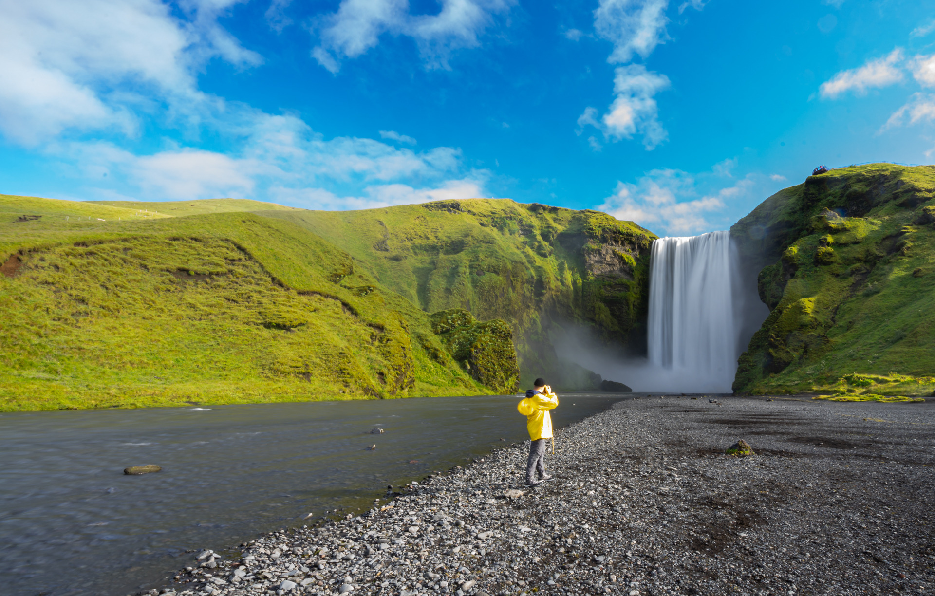 Osoba w żółtej kurtce fotografuje wodospad Skógafoss w Islandii, z zielonymi wzgórzami i błękitnym niebem z chmurami w tle.