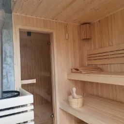 Wnętrze sauny wykończone jasnym drewnem, z piecem na drewno i kominem ze stali nierdzewnej, drewnianymi ławkami na różnych poziomach oraz drewnianym czerpakiem i wiadrem na dolnej ławce.