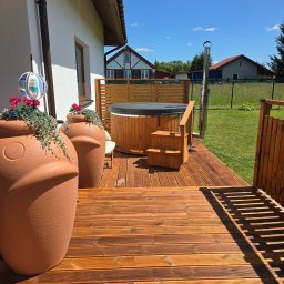 WISDOM - Drewniany taras z jacuzzi i dużymi donicami w kształcie głowy. Elementy drewniane w ciepłym odcieniu brązu. W tle domy i błękitne niebo.