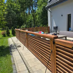 Drewniane, lamelowe ogrodzenie tarasu z ozdobnymi lampionami, wsparte na metalowych podporach. W tle dom z szarą elewacją i zieleń drzew.