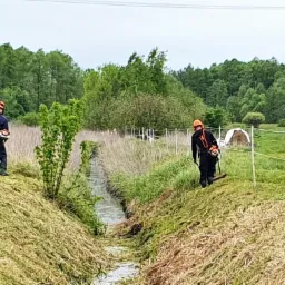 Dwaj pracownicy w kombinezonach i kaskach koszą trawę kosiarkami spalinowymi wzdłuż rowu melioracyjnego na tle drzew i pastwiska z koniem.