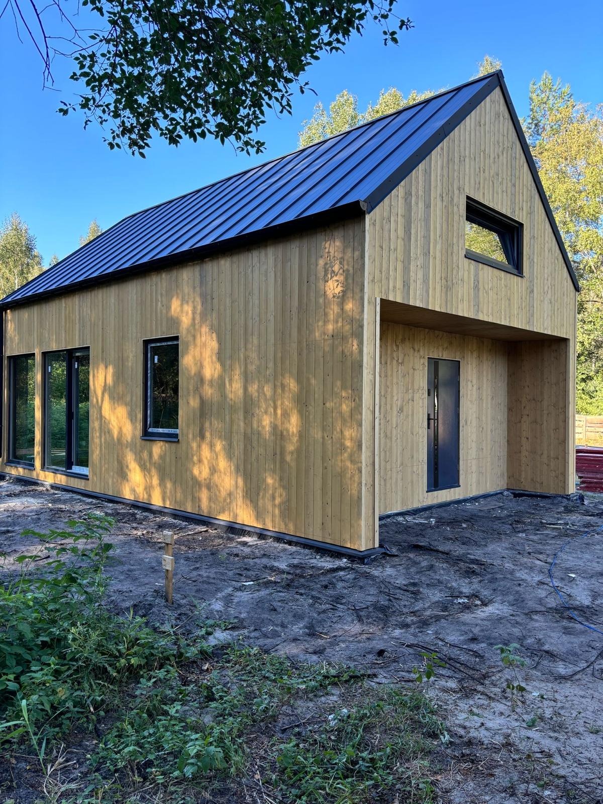 Nowoczesny dom z drewnianą elewacją i ciemnym dachem blachodachówką, duże okna z czarnymi ramami, widoczne ślady prac ziemnych wokół budynku.