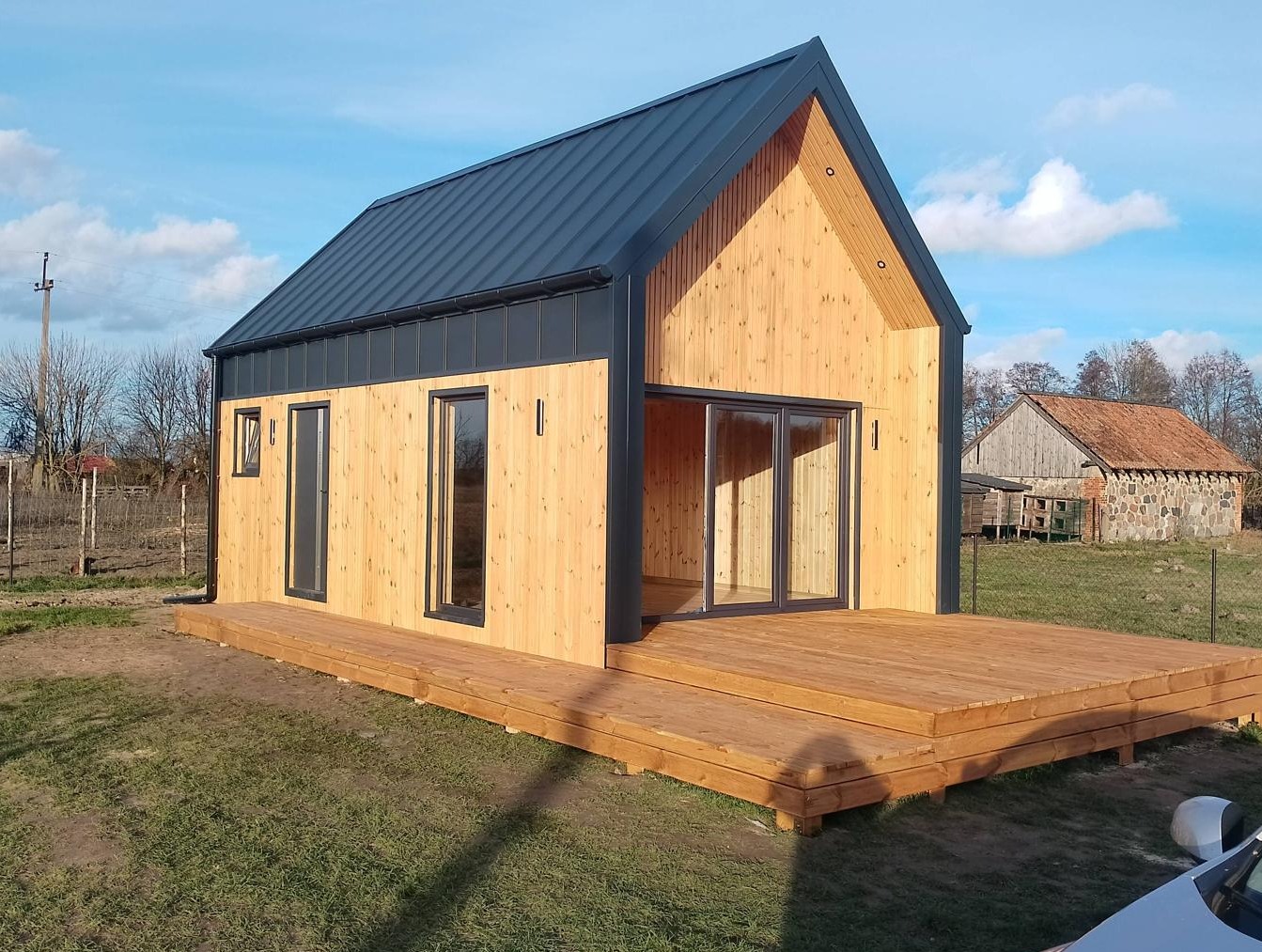 Nowoczesny drewniany domek z ciemną blachodachówką i tarasem z desek, widok na wiejski krajobraz z budynkiem gospodarczym w tle.