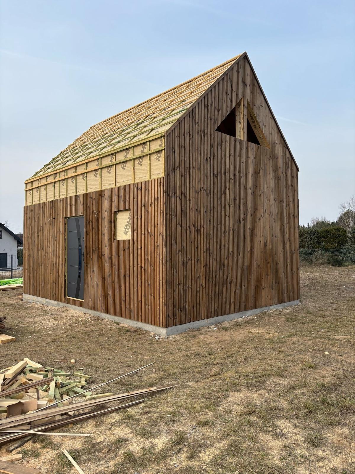 Budowa nowoczesnej, drewnianej konstrukcji z pionowymi deskami, widoczna izolacja dachu i otwór okienny z naklejką, na ziemi leżą resztki drewna.